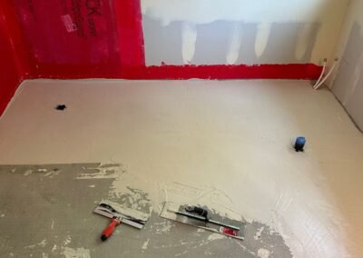 A prepared bathroom floor with tools for leveling and smoothing the surface, ready for tile installation. Red waterproofing material is visible on the lower wall, ensuring the area is prepped for the next steps of the renovation process.