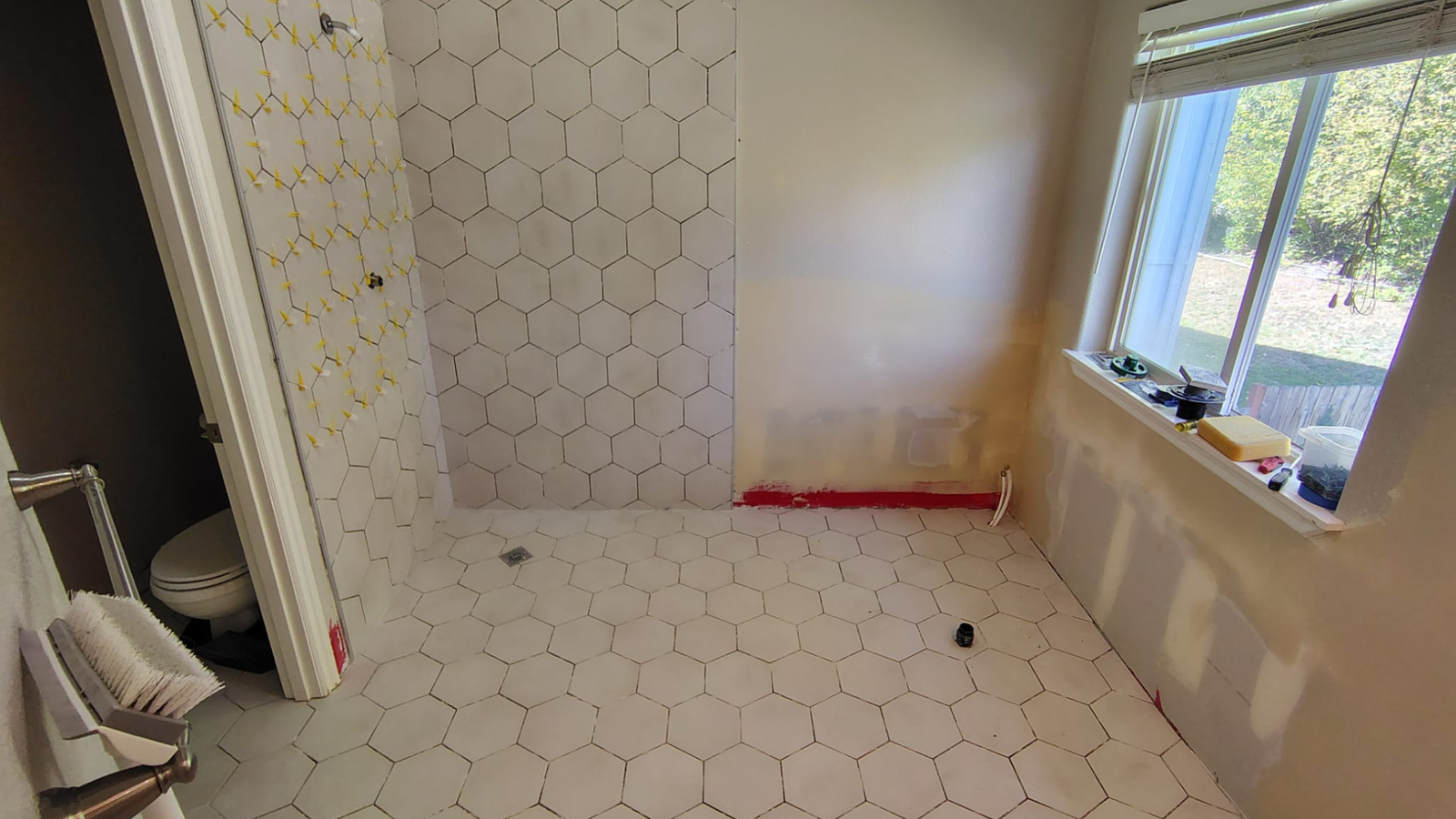 Close-up view of a hexagonal tile floor in a bathroom before grout application. The tiles are evenly spaced, and the preparation phase is visible, with unfinished areas showing where grout will be applied. Light from a nearby window casts shadows across the clean, new tile surface, emphasizing the hexagonal pattern.