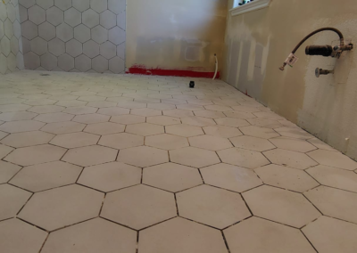 Close-up view of a hexagonal tile floor in a bathroom before grout application. The tiles are evenly spaced, and the preparation phase is visible, with unfinished areas showing where grout will be applied. Light from a nearby window casts shadows across the clean, new tile surface, emphasizing the hexagonal pattern.