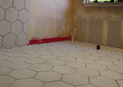 Close-up view of a hexagonal tile floor in a bathroom before grout application. The tiles are evenly spaced, and the preparation phase is visible, with unfinished areas showing where grout will be applied. Light from a nearby window casts shadows across the clean, new tile surface, emphasizing the hexagonal pattern.