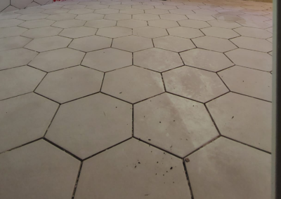 Close-up view of a hexagonal tile floor in a bathroom before grout application. The tiles are evenly spaced, and the preparation phase is visible, with unfinished areas showing where grout will be applied. Light from a nearby window casts shadows across the clean, new tile surface, emphasizing the hexagonal pattern.